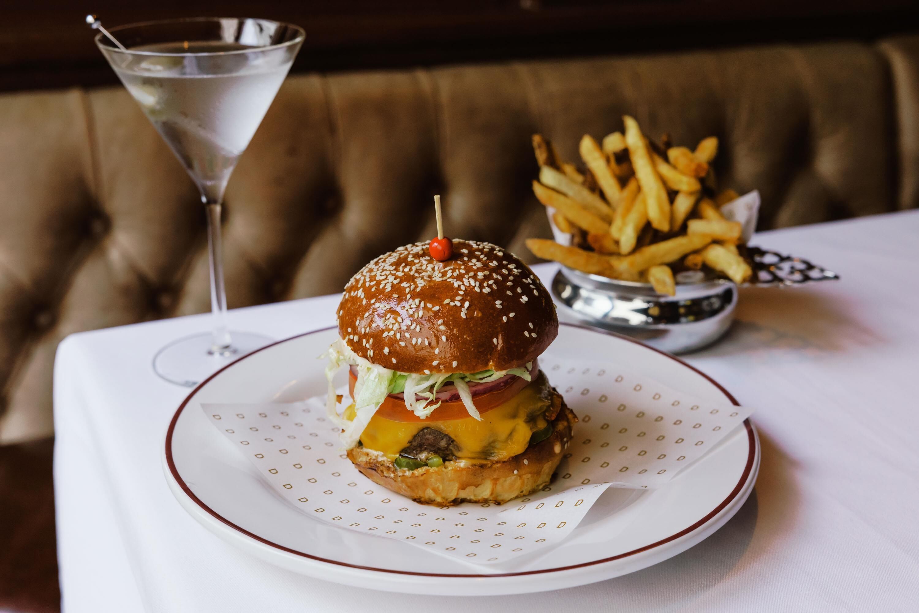 Un hamburger accompagné de frites, servi sur une assiette blanche, un verre d’eau et, au fond, un bol rempli de frites.