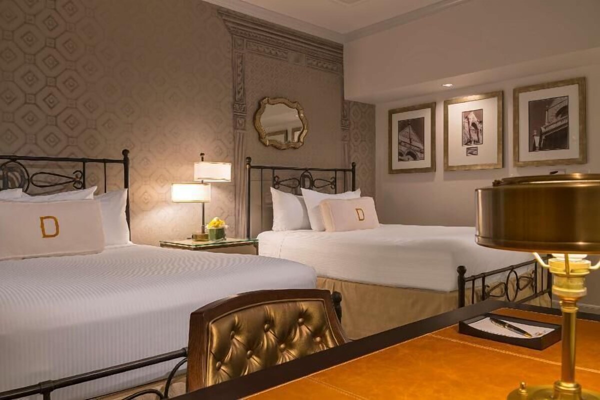 A hotel room with two beds, a mirror, framed pictures on the wall, and a table with a lamp.