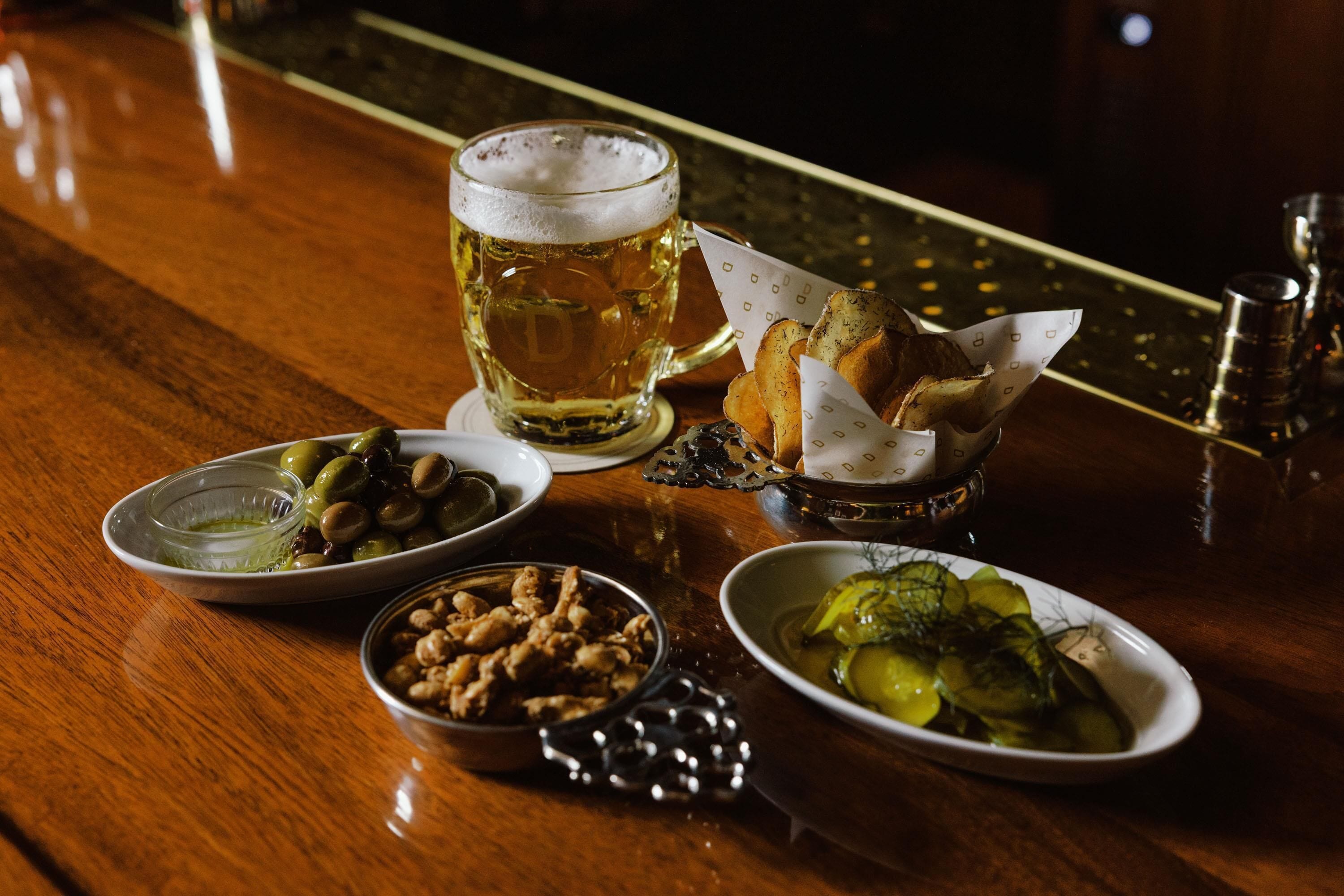 A wooden table with a glass of beer, bread, olives, nuts, and pickles.