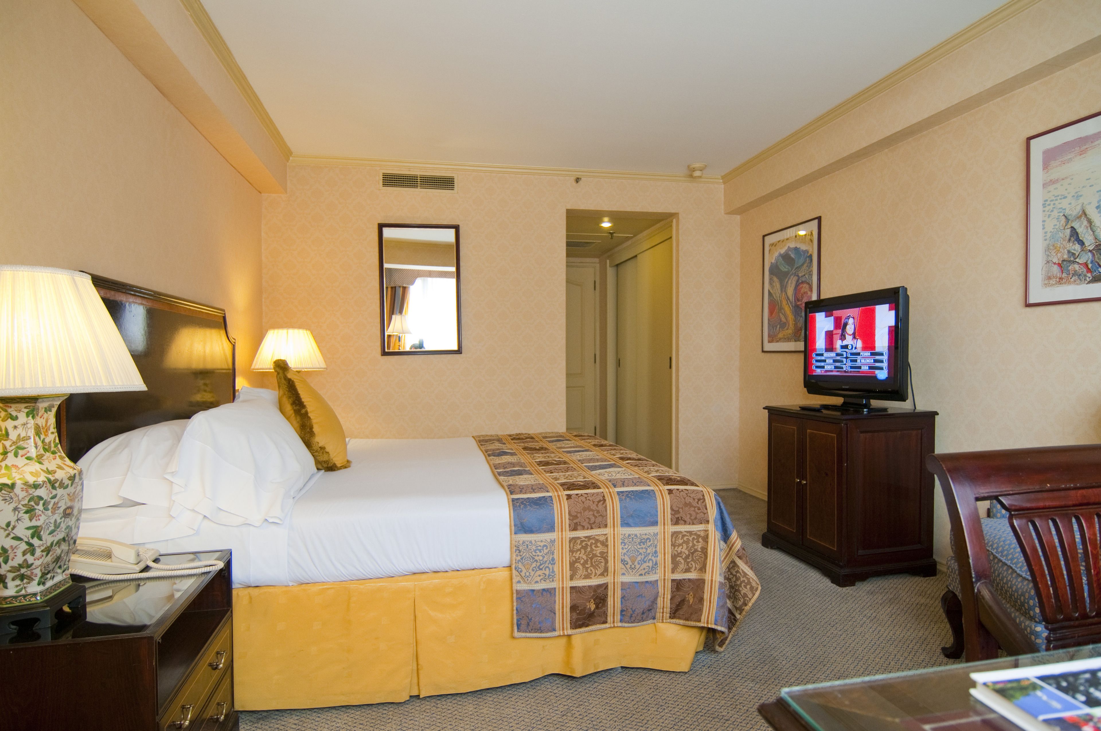 A hotel room with a bed, a television, a lamp, a mirror, and a door leading to another room.