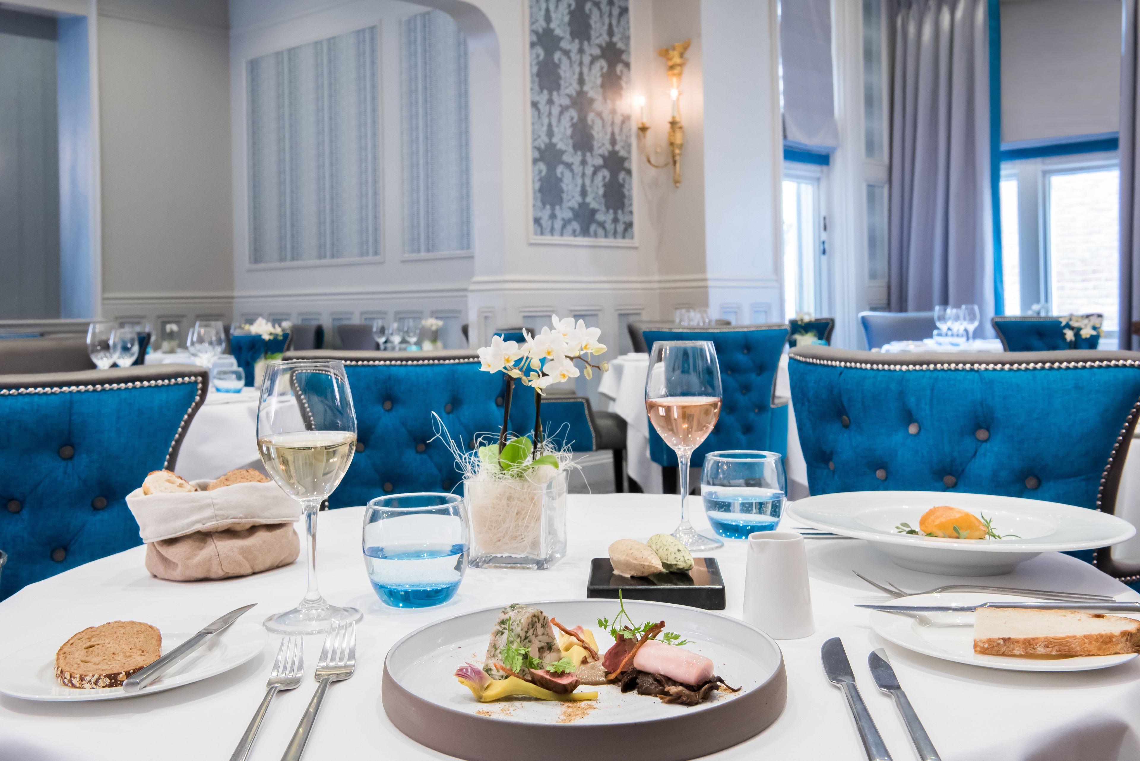 A dining table set with plates of food, glasses, and cutlery in a room with blue upholstered chairs and a decorative wall.