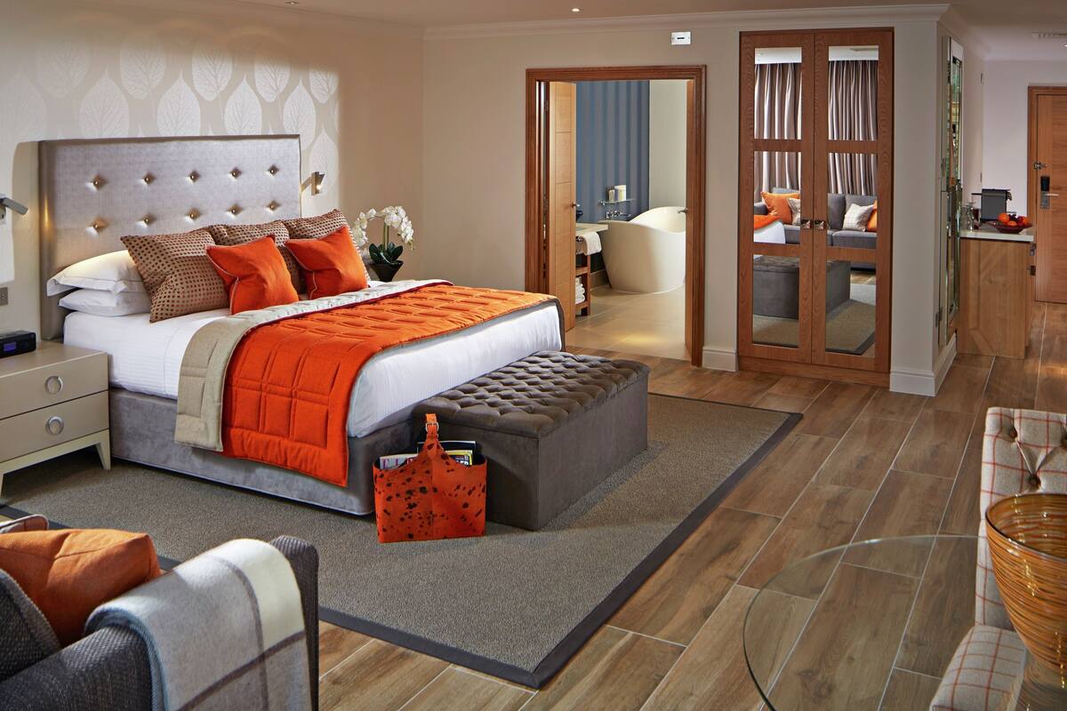 A modern bedroom with a large bed, a grey ottoman, a bedside table, and a small table with a glass top.