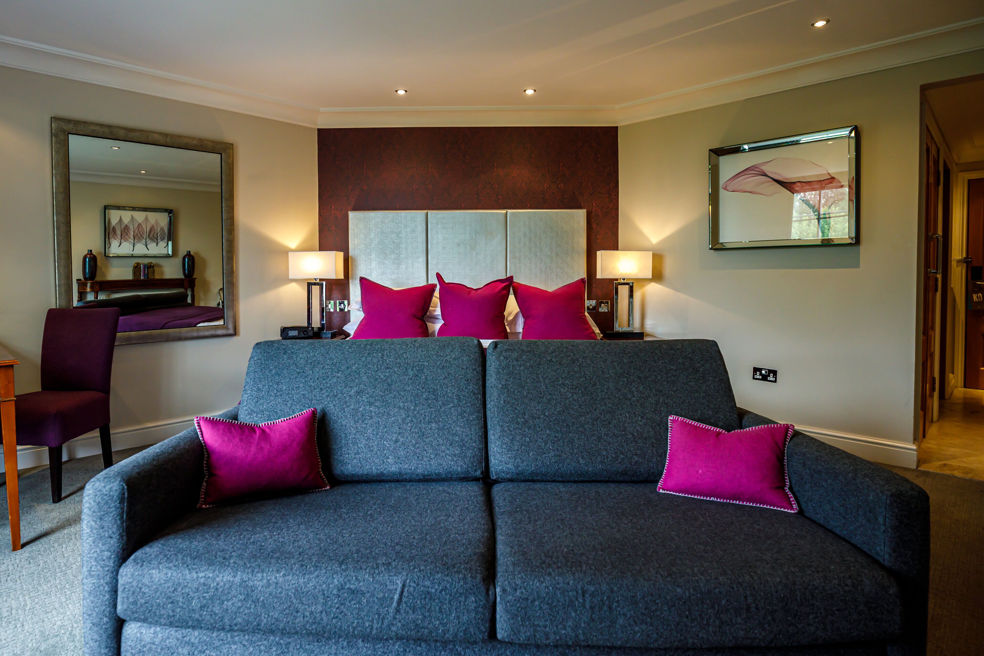 A modern hotel room with a grey sofa, two purple pillows, a large mirror, and a framed artwork on the wall.