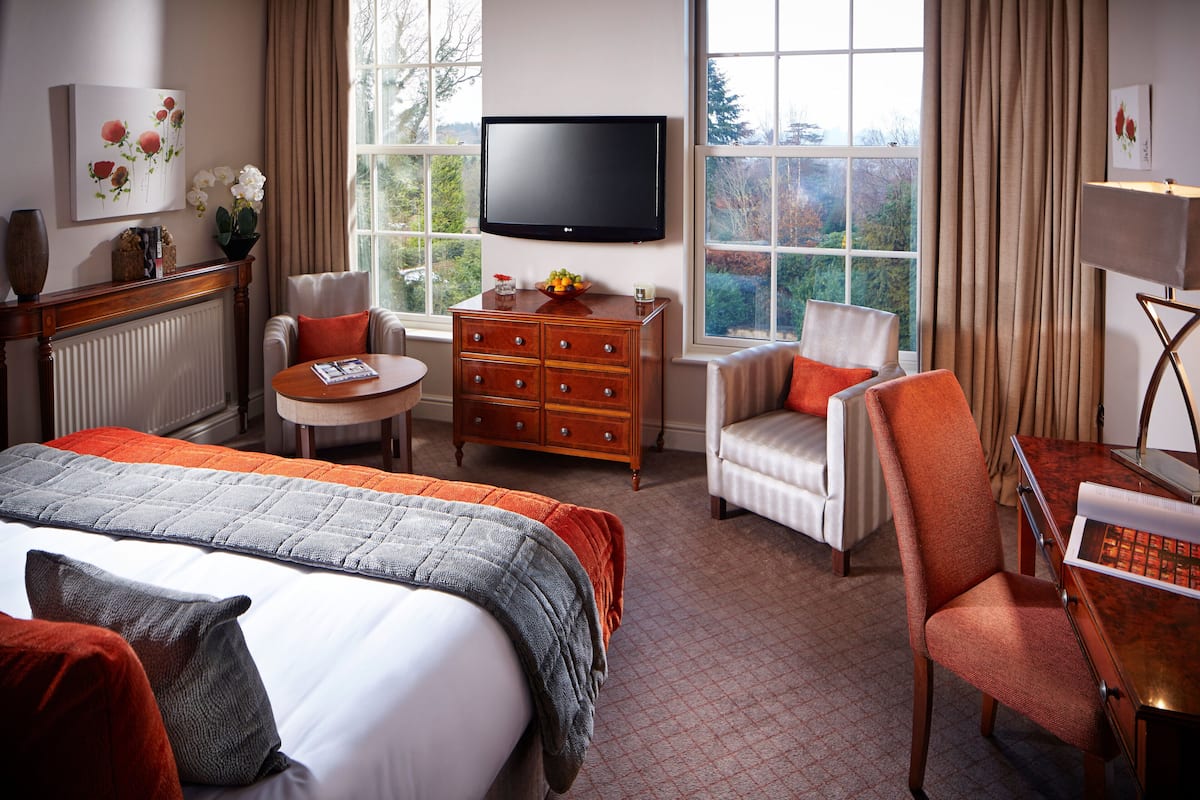 A hotel room with a large bed, two armchairs, a wooden dresser, a television, and a view of the outdoors.