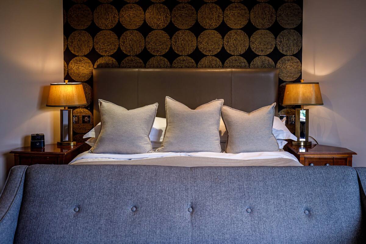 A neatly made bed with a grey headboard, two bedside lamps, and a patterned wall behind.