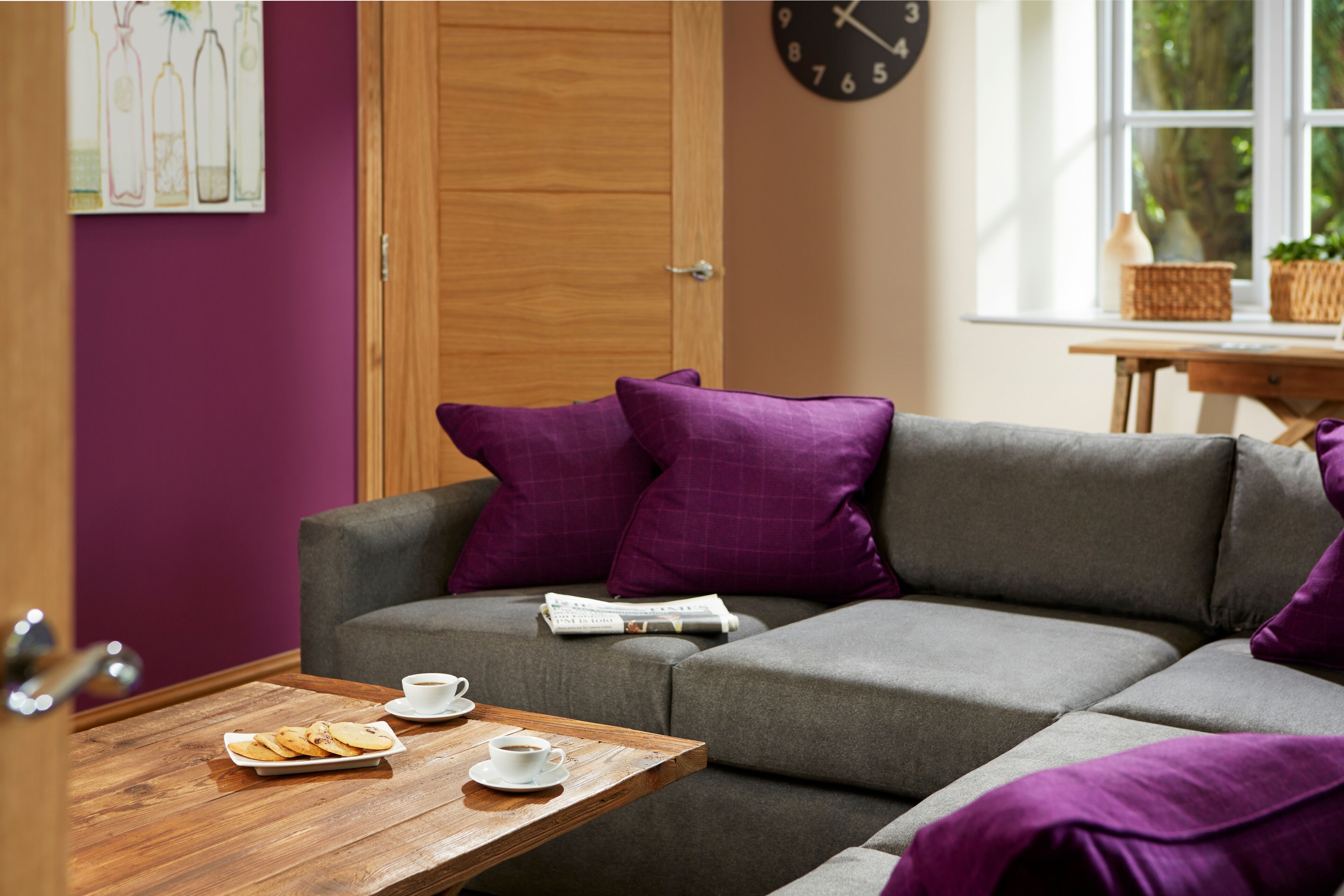 Un salón moderno con un sofá gris, cojines morados, una mesa de centro de madera con pasteles y una taza, y un reloj en la pared.