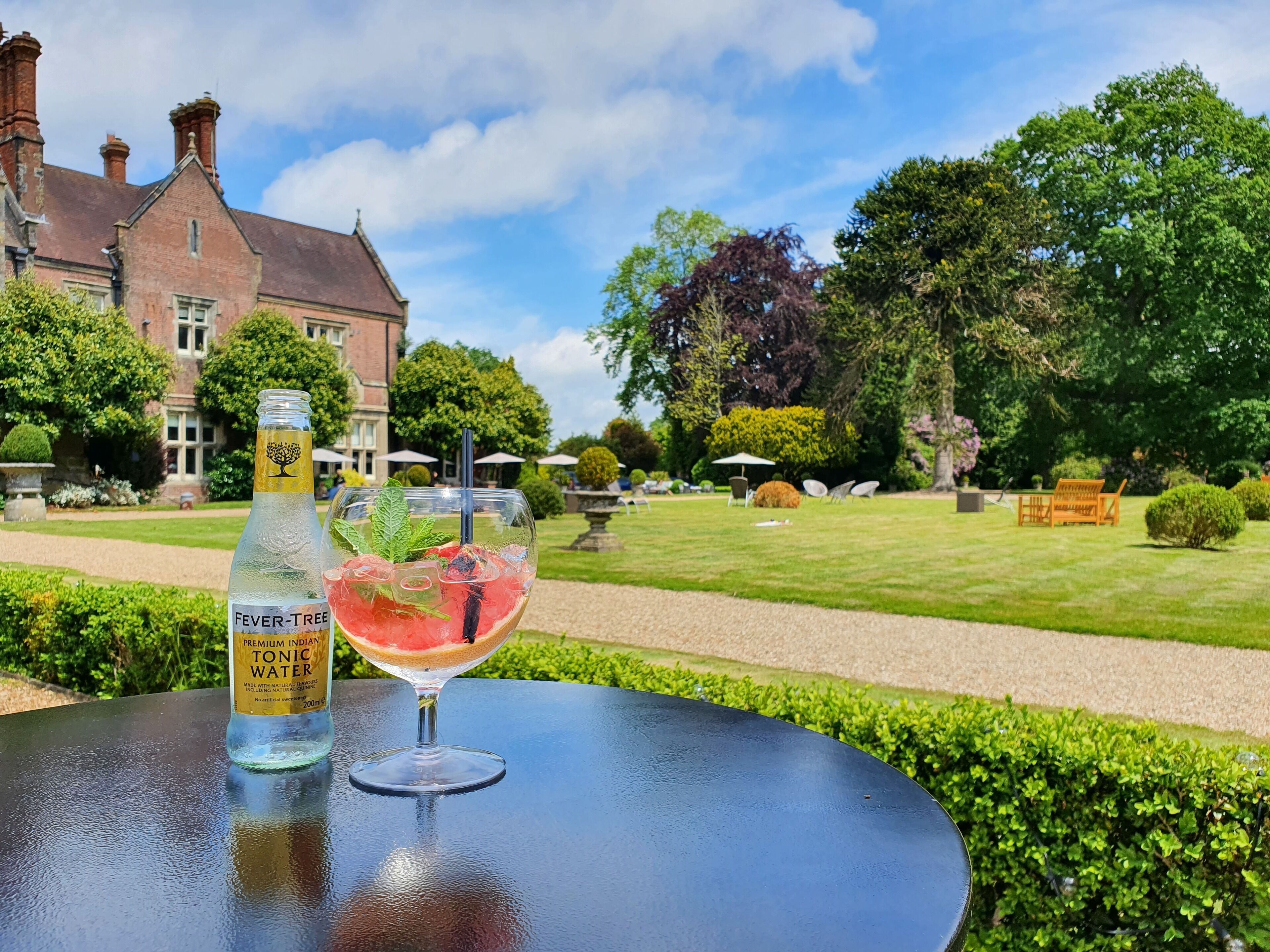 Una botella de Fever Tree Premium Tónica India y un vaso con una rodaja de sandía sobre una mesa en un jardín.