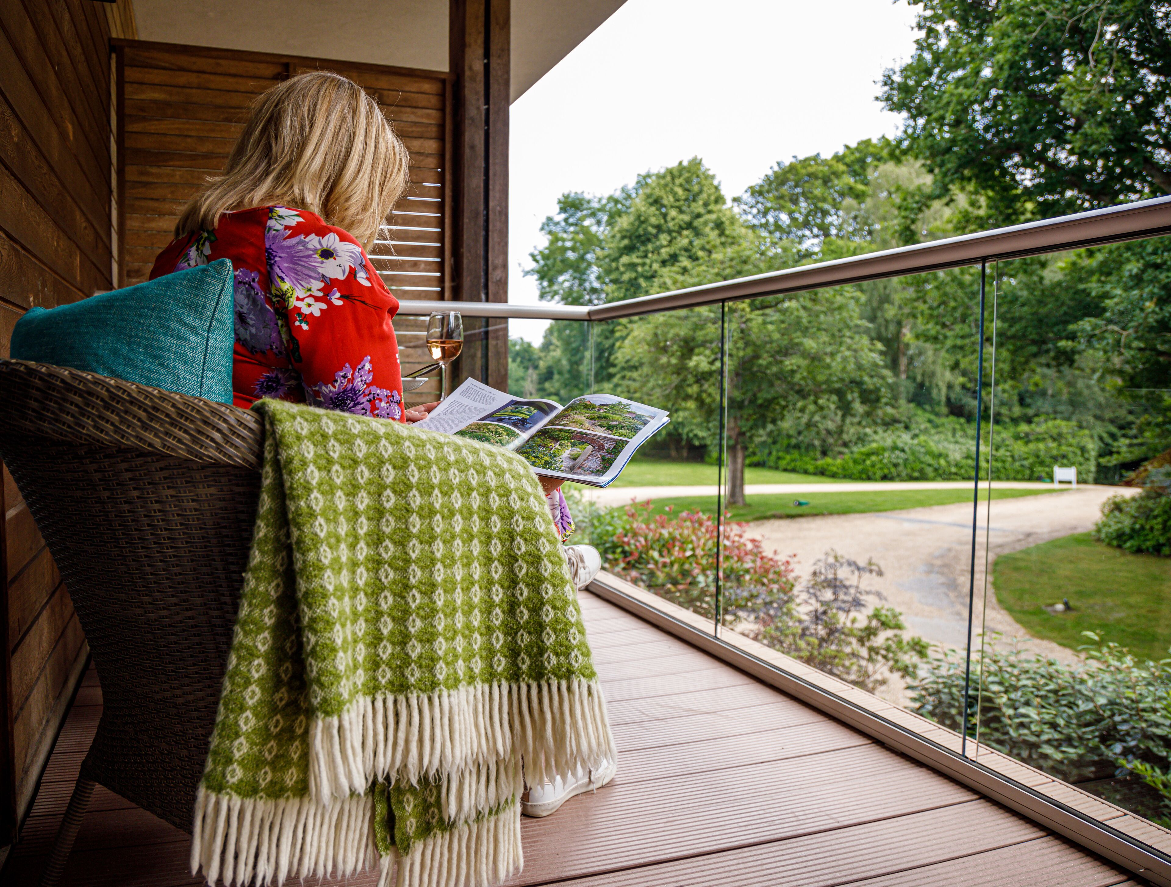 Une personne est assise sur un balcon, un livre à la main, et profite de la vue sur un jardin et un sentier.