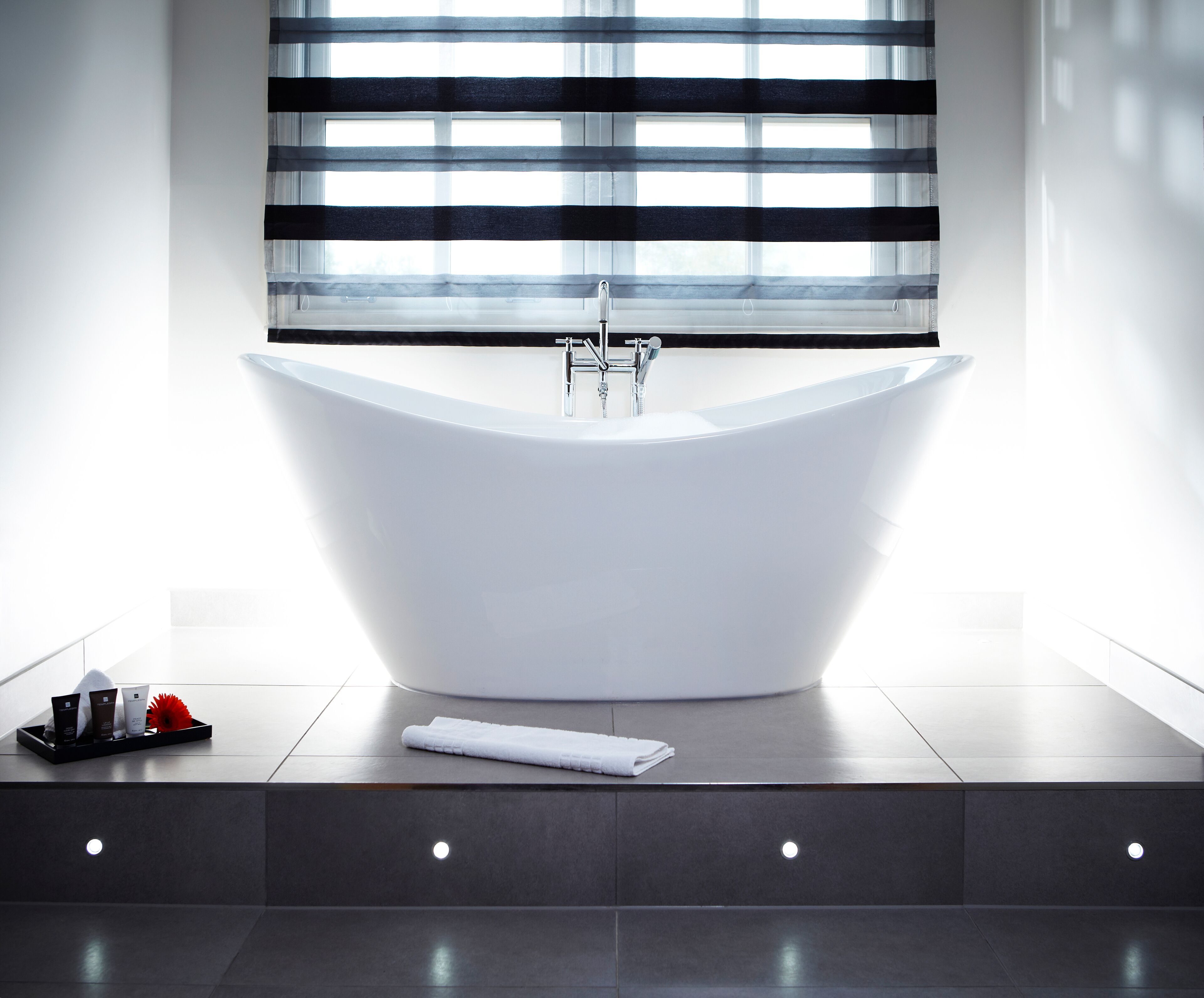 Ein modernes Badezimmer mit freistehender Badewanne, einem Fenster mit horizontalen Jalousien und einer Ablage mit Toilettenartikeln.