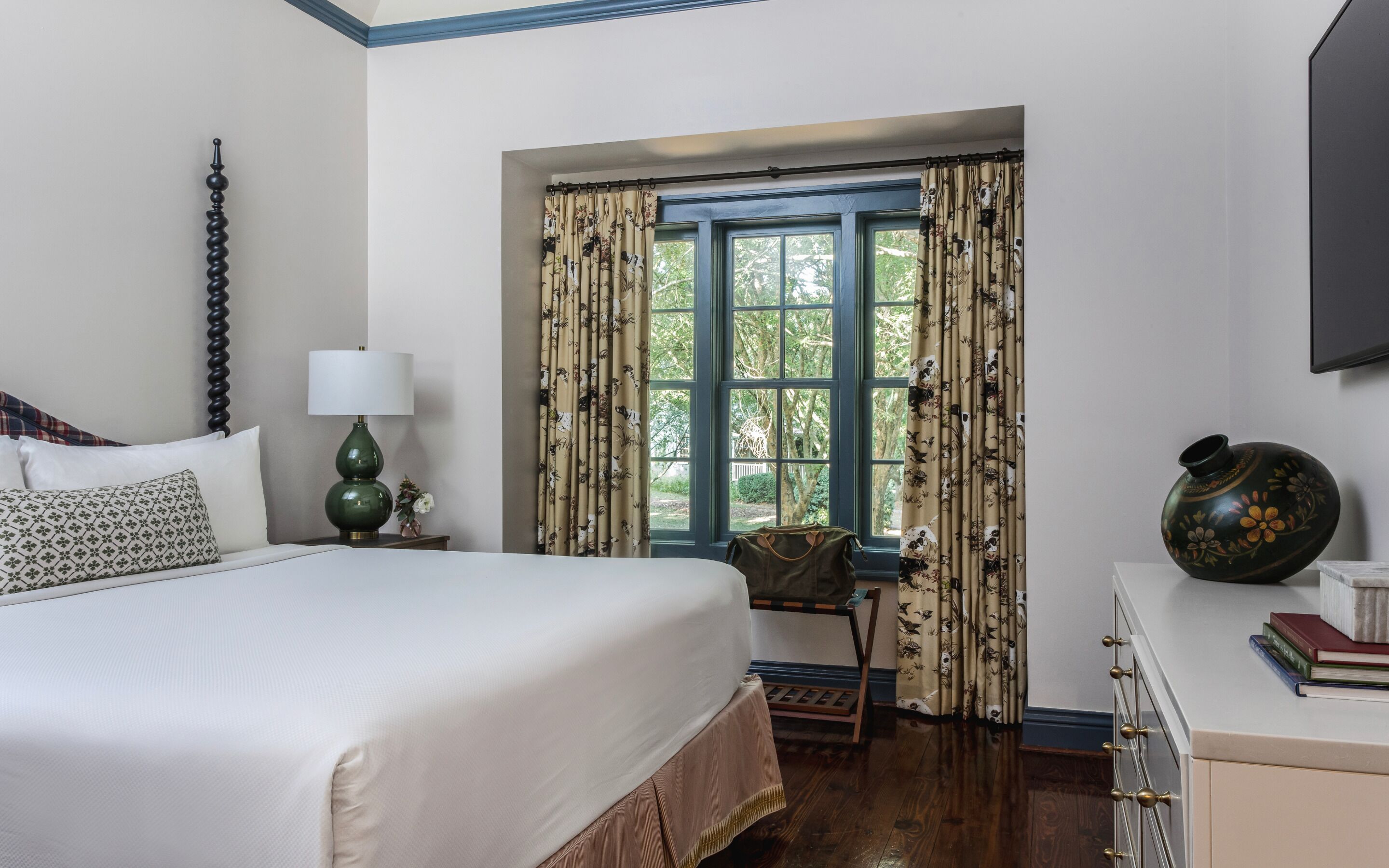 A bedroom with a bed, a nightstand with a lamp, a dresser, a window with curtains, and a vase on the dresser.