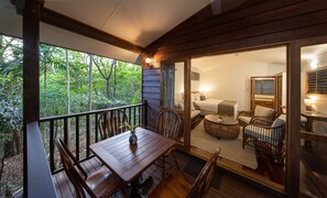 A wooden deck with a table and chairs, a bedroom with a bed and seating area, and a view of the surrounding forest. - Thala Beach Nature Reserve (Oak Beach)