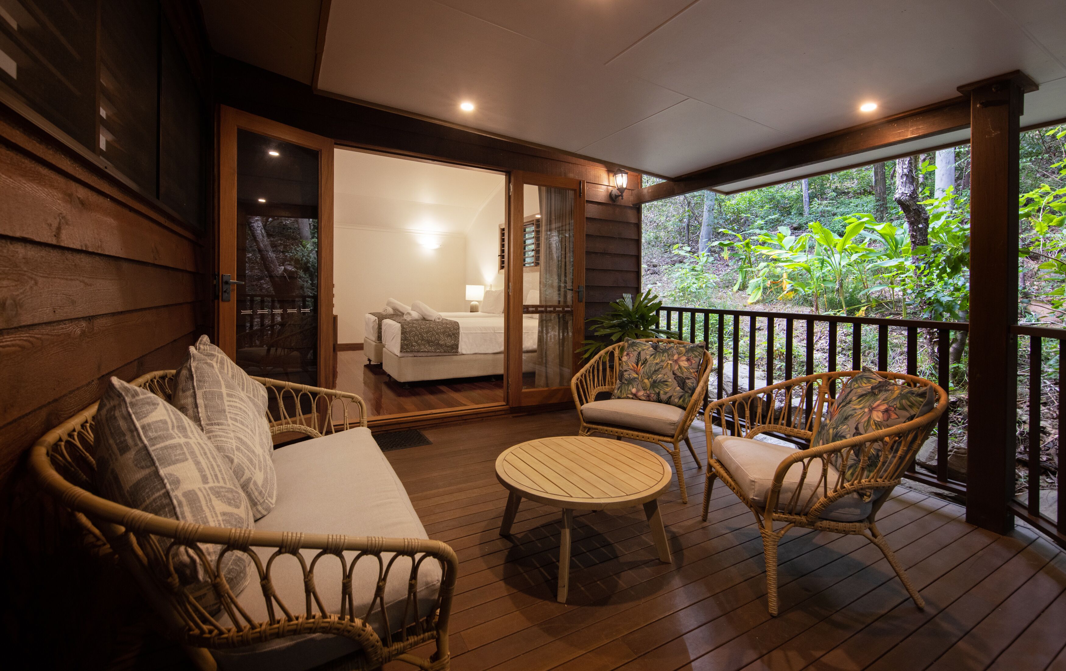 A wooden deck with wicker furniture, a glass door leading to a bedroom, and a view of greenery.