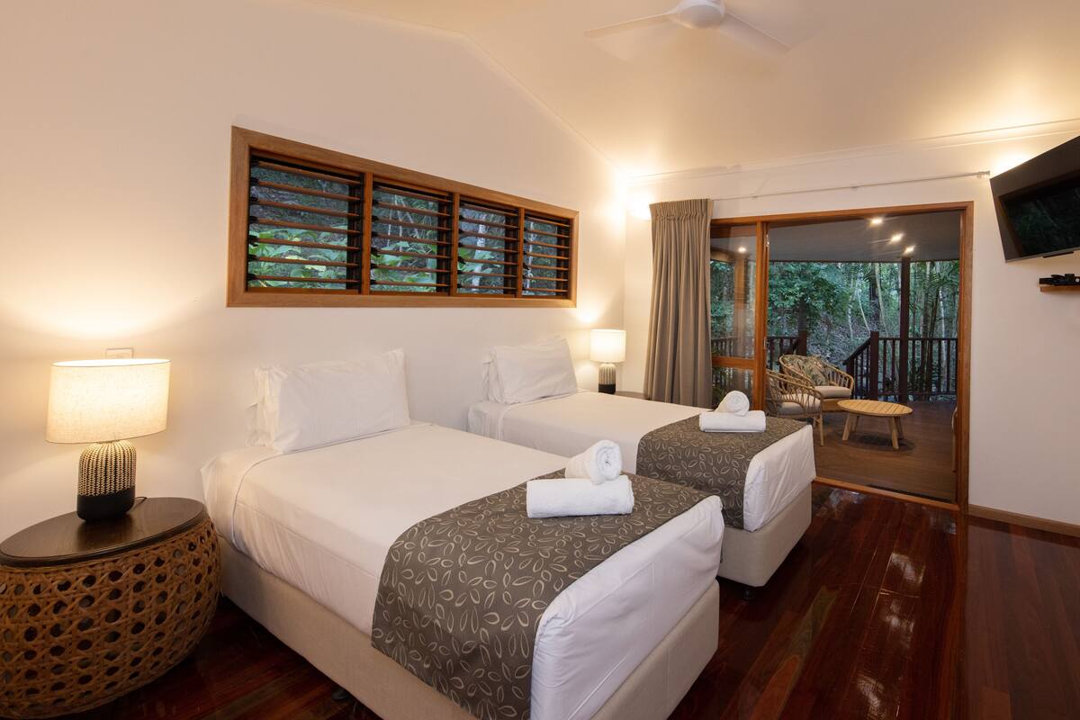 A hotel room with two beds, a TV, a balcony with a view of trees, and a wooden floor.