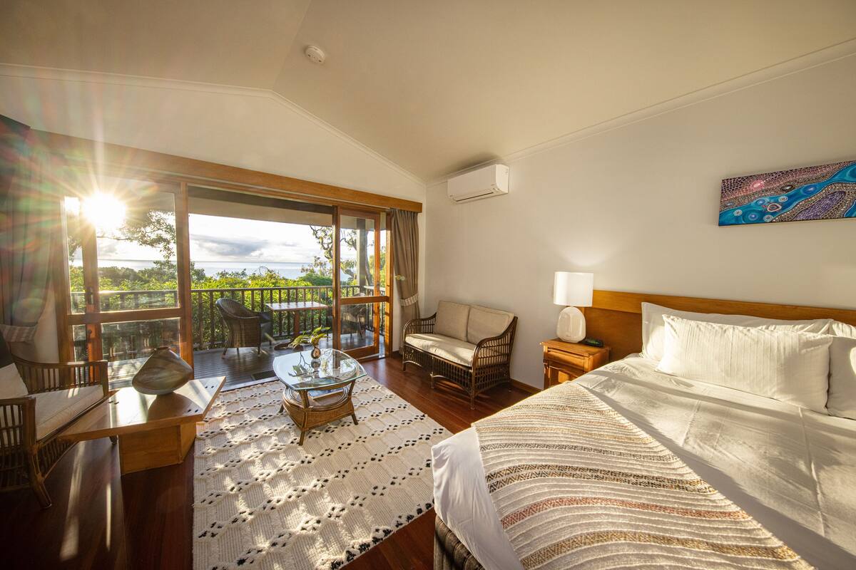 A hotel room with a large bed, a glass table, wicker chairs, and a view of the outdoors.