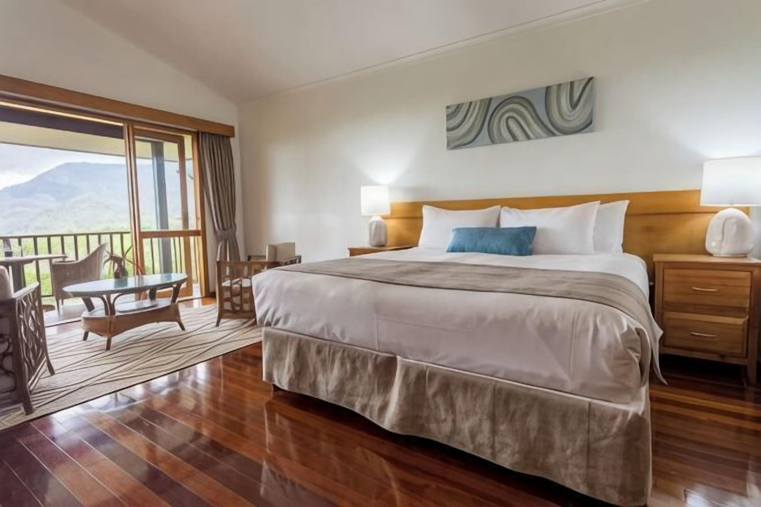Quarto de hotel com uma cama grande, uma varanda com mesa e cadeiras, e vista para as montanhas.