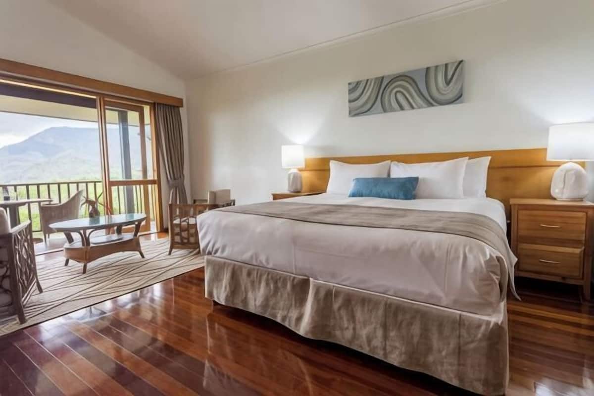 Quarto de hotel com uma cama grande, uma varanda com mesa e cadeiras, e vista para as montanhas.