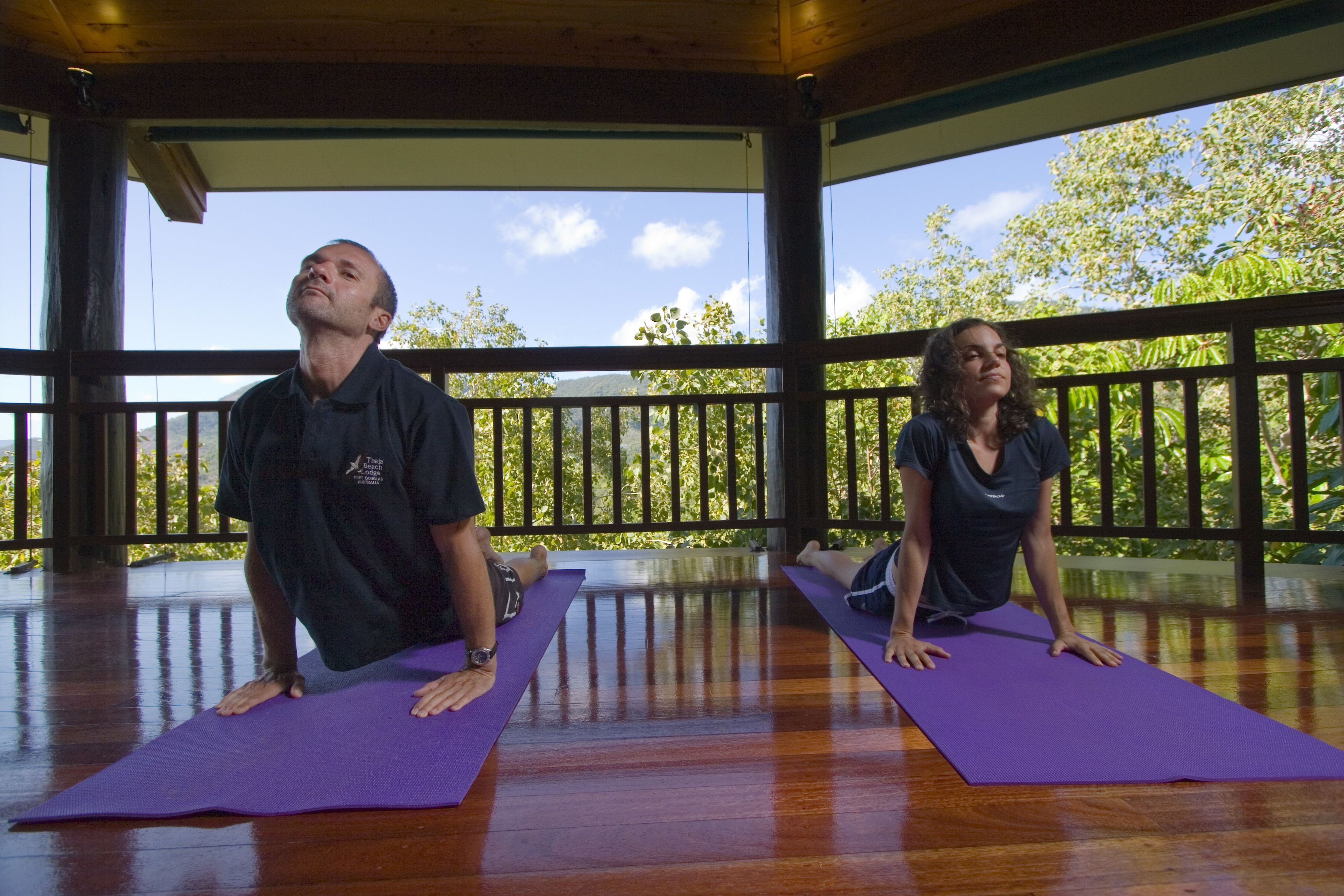 Two people on purple yoga mats in a wooden-floored room with a balcony.