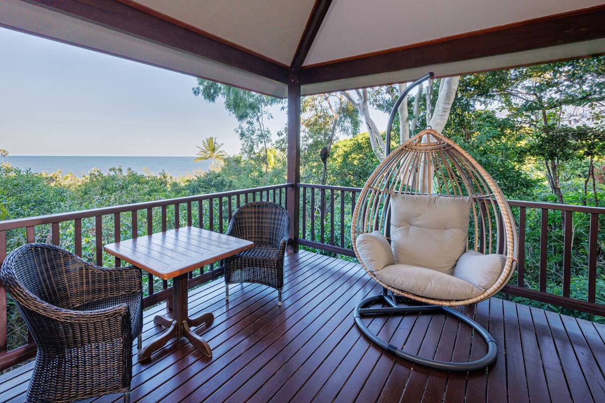 A wooden deck with wicker furniture, a hanging chair, and a table overlooking a lush landscape and the sea.