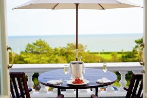 A round table with a white umbrella, a silver pitcher, and two glasses of white wine, set on a balcony with a view of the ocean.