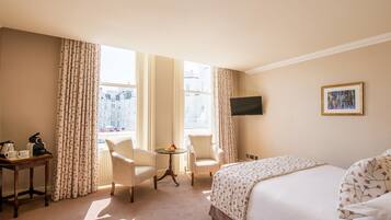 A hotel room with a bed, two armchairs, a small table with a fruit bowl, a coffee maker, and a TV on the wall.