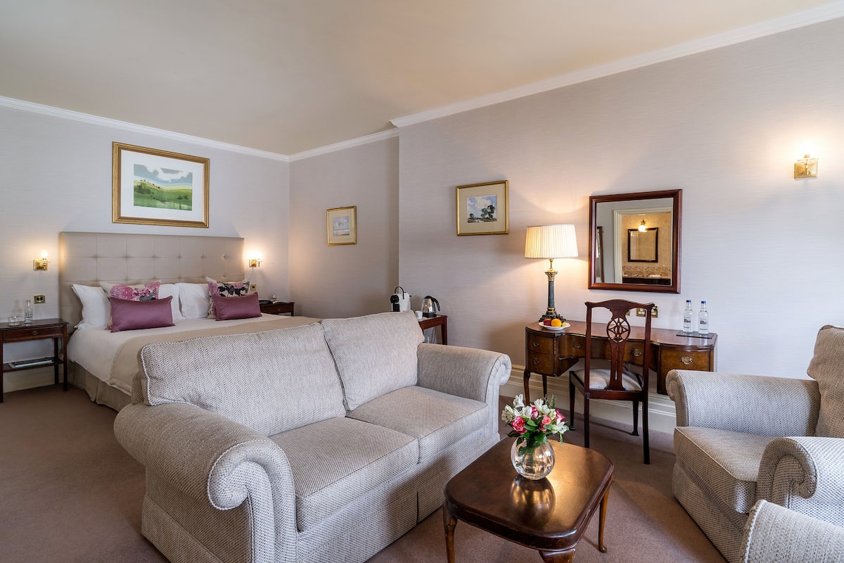 Habitación de hotel con cama, dos sofás, una mesita de centro, un escritorio y una silla. Se observan cuadros enmarcados en las paredes y una lámpara sobre el escritorio.