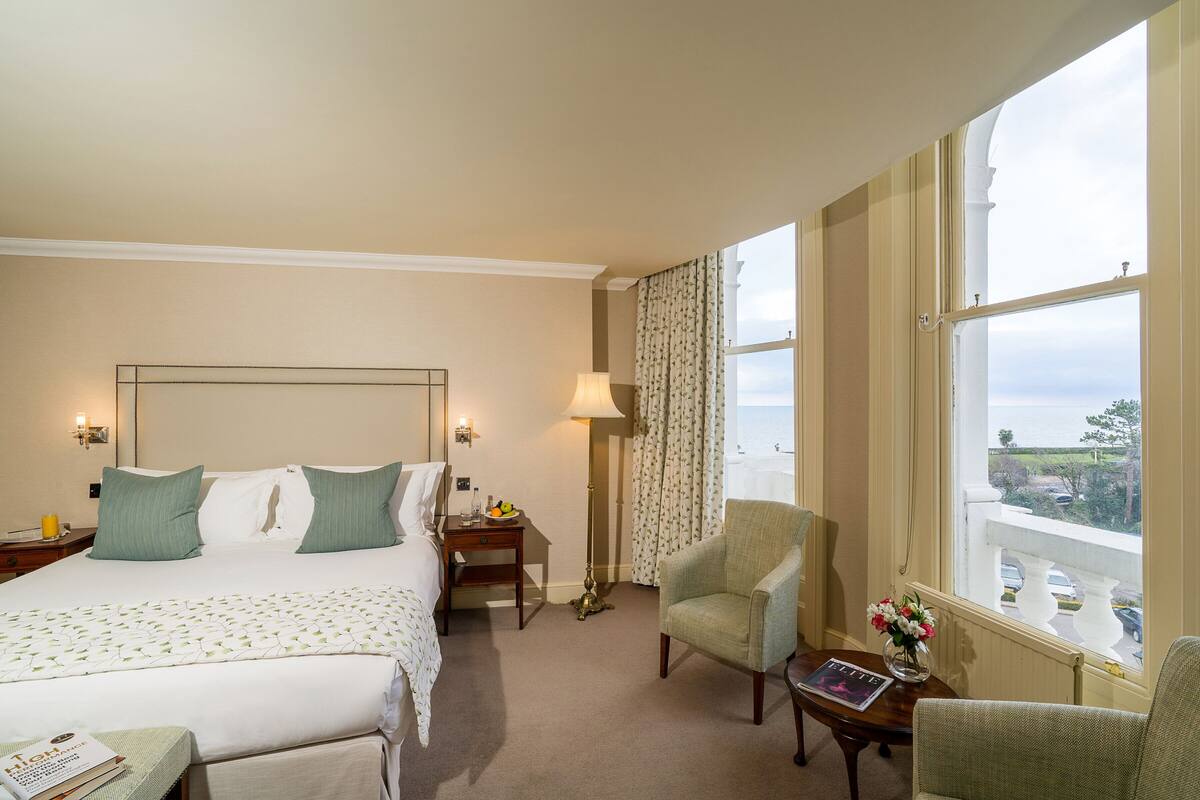 Habitación de hotel con una cama grande, un área de descanso con una silla y una mesa, una lámpara y vistas al exterior a través de una ventana.