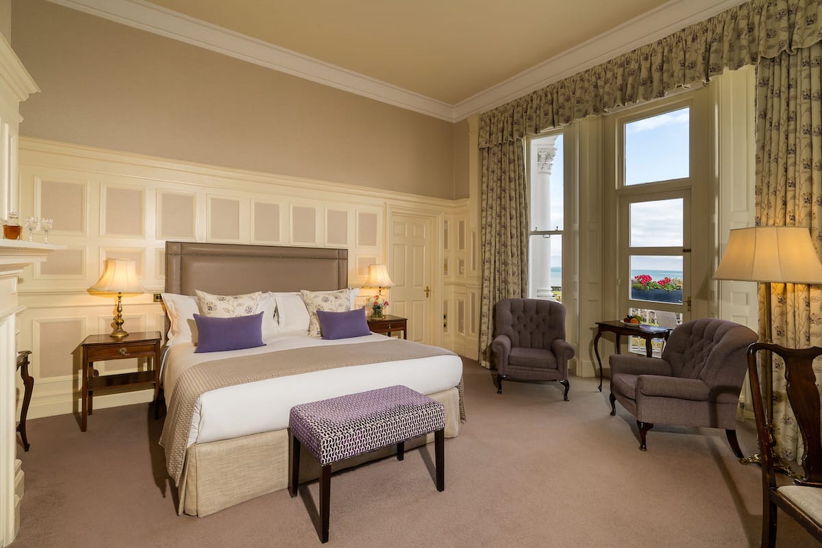 Habitación de hotel con una cama grande, dos mesitas de noche, un banco, dos sillones, una mesita pequeña y vistas al exterior.