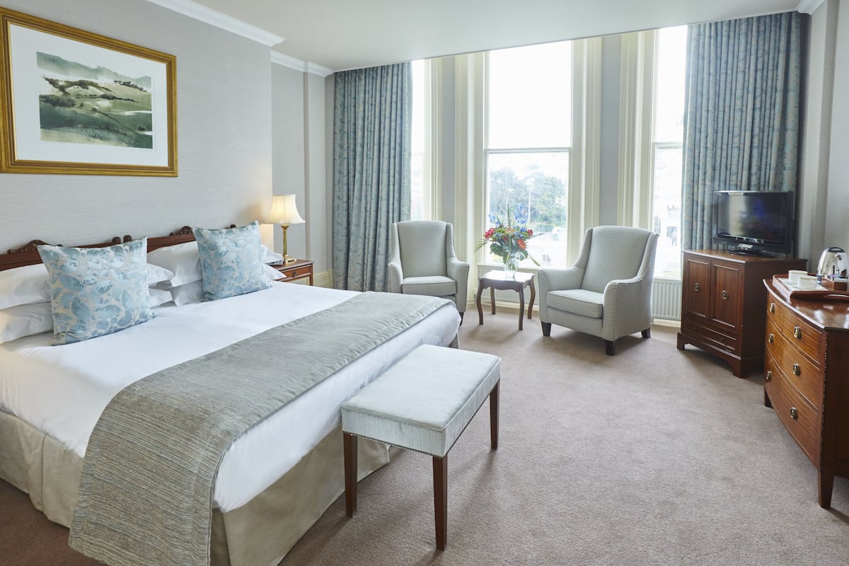 Habitación de hotel con una cama grande, dos sillones, una mesita con flores, un televisor y un aparador.