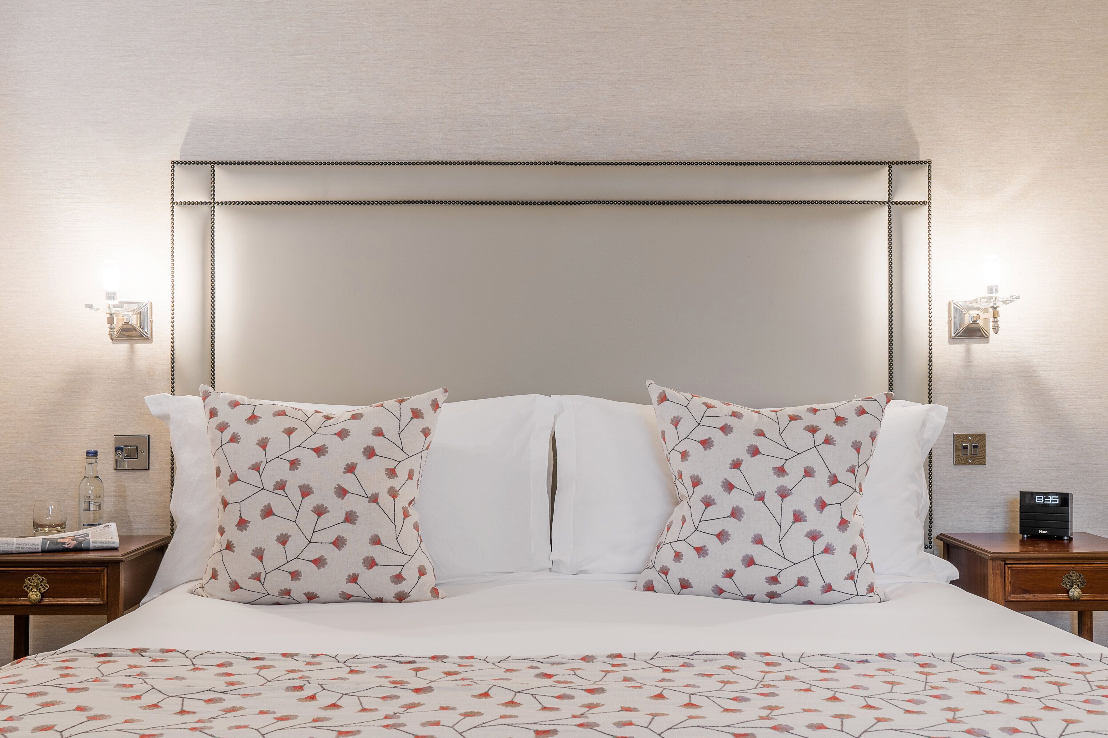 A neatly made bed with floral-patterned pillows and a white headboard. There are two bedside tables, one with a bottle and a glass, and the other with a clock.
