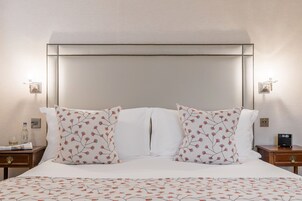 A neatly made bed with floral-patterned pillows and a white headboard. There are two bedside tables, one with a bottle and a glass, and the other with a clock.