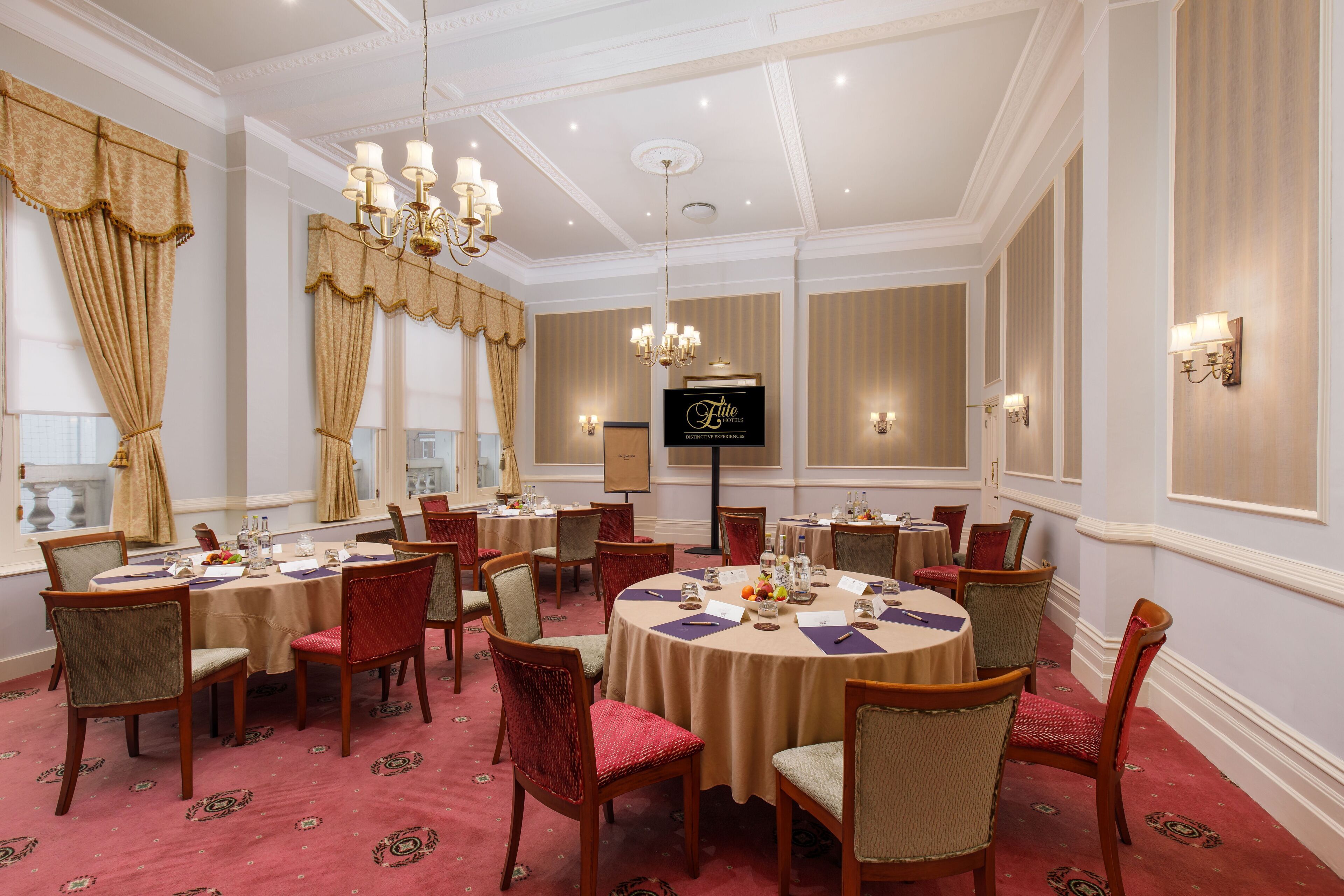 A formal dining room with round tables, red upholstered chairs, and a chandelier.