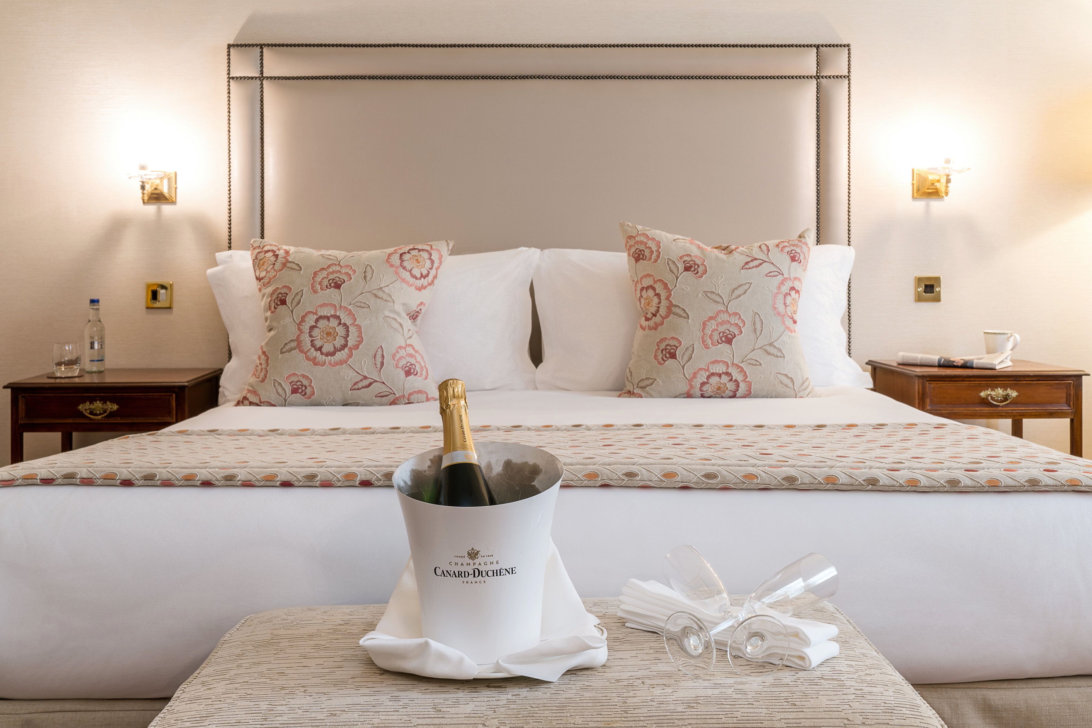 Un lit bien fait, agrémenté de coussins à motifs floraux, un seau à champagne et une serviette pliée posés sur une table de chevet.