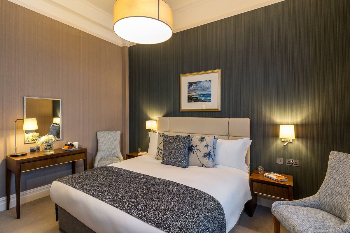Habitación de hotel con una cama grande, una mesita de noche con lámpara, una silla y un cuadro colgado en la pared.