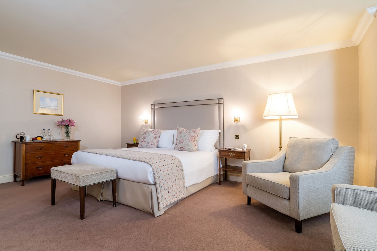 Habitación de hotel con una cama grande, una mesita de noche, una silla, un tocador y una lámpara.