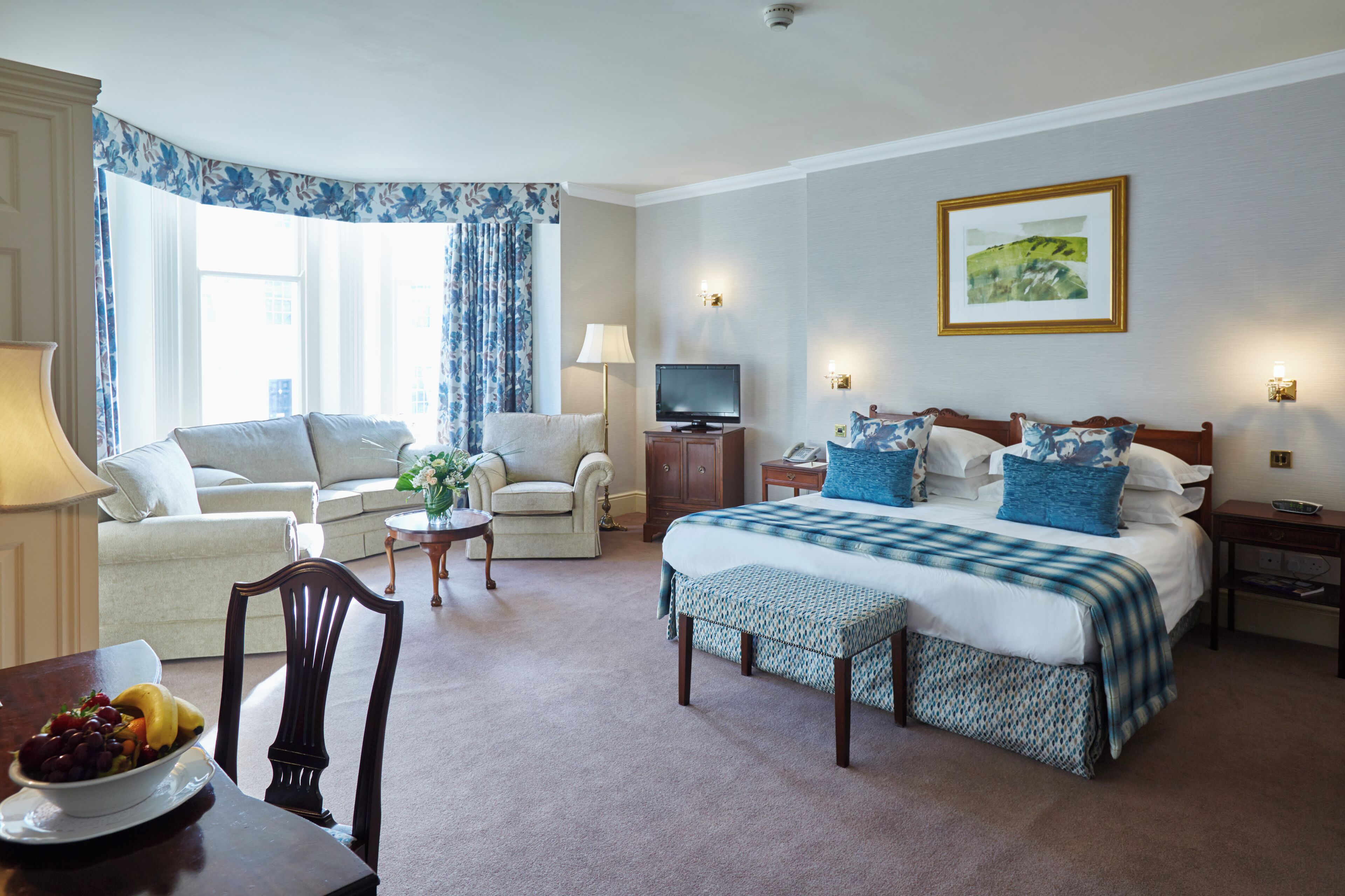 A hotel room with a bed, a sofa, a TV, and a dining table with fruit.