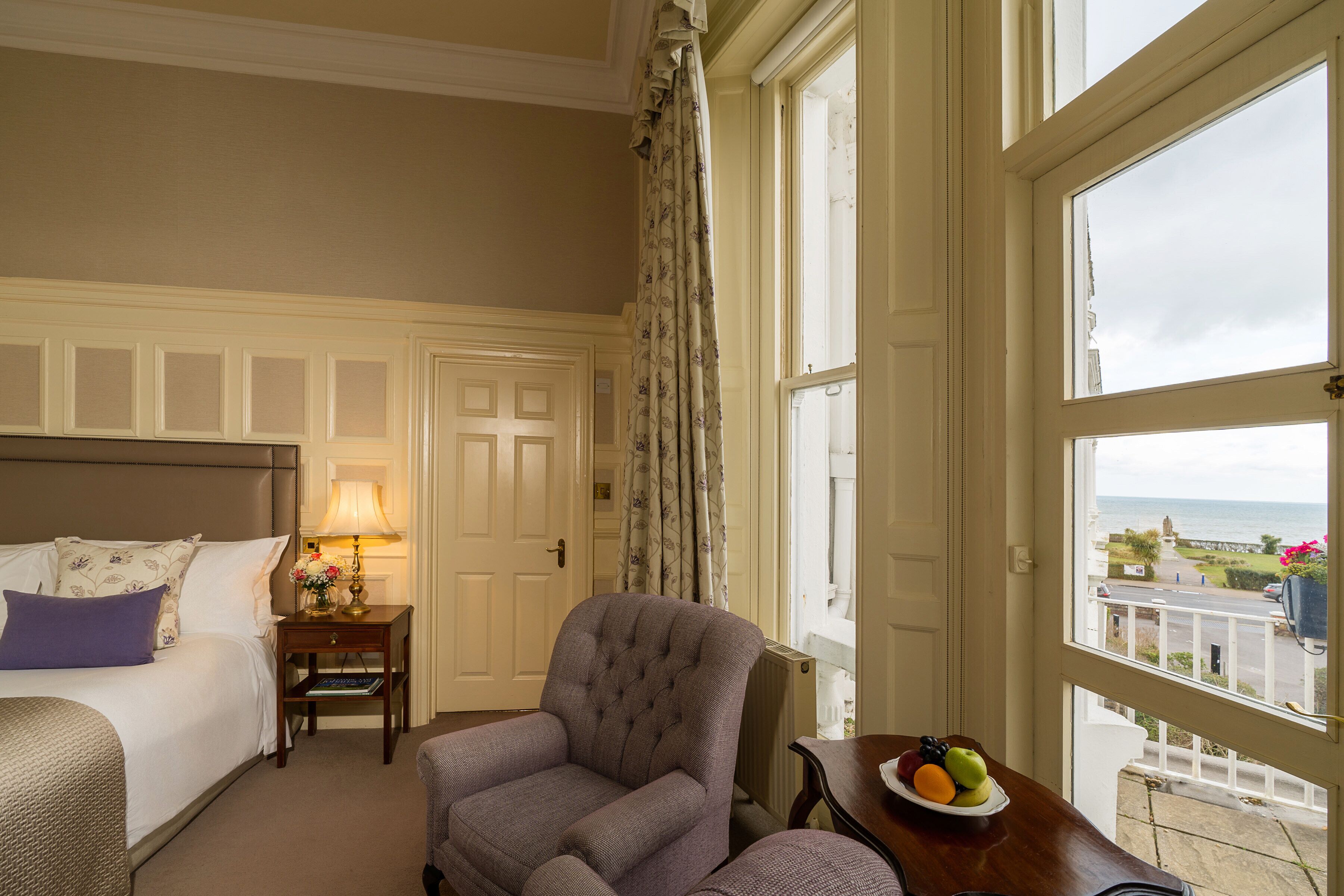 A hotel room with a bed, a chair, a small table with a fruit bowl, and a view of the sea through large windows.