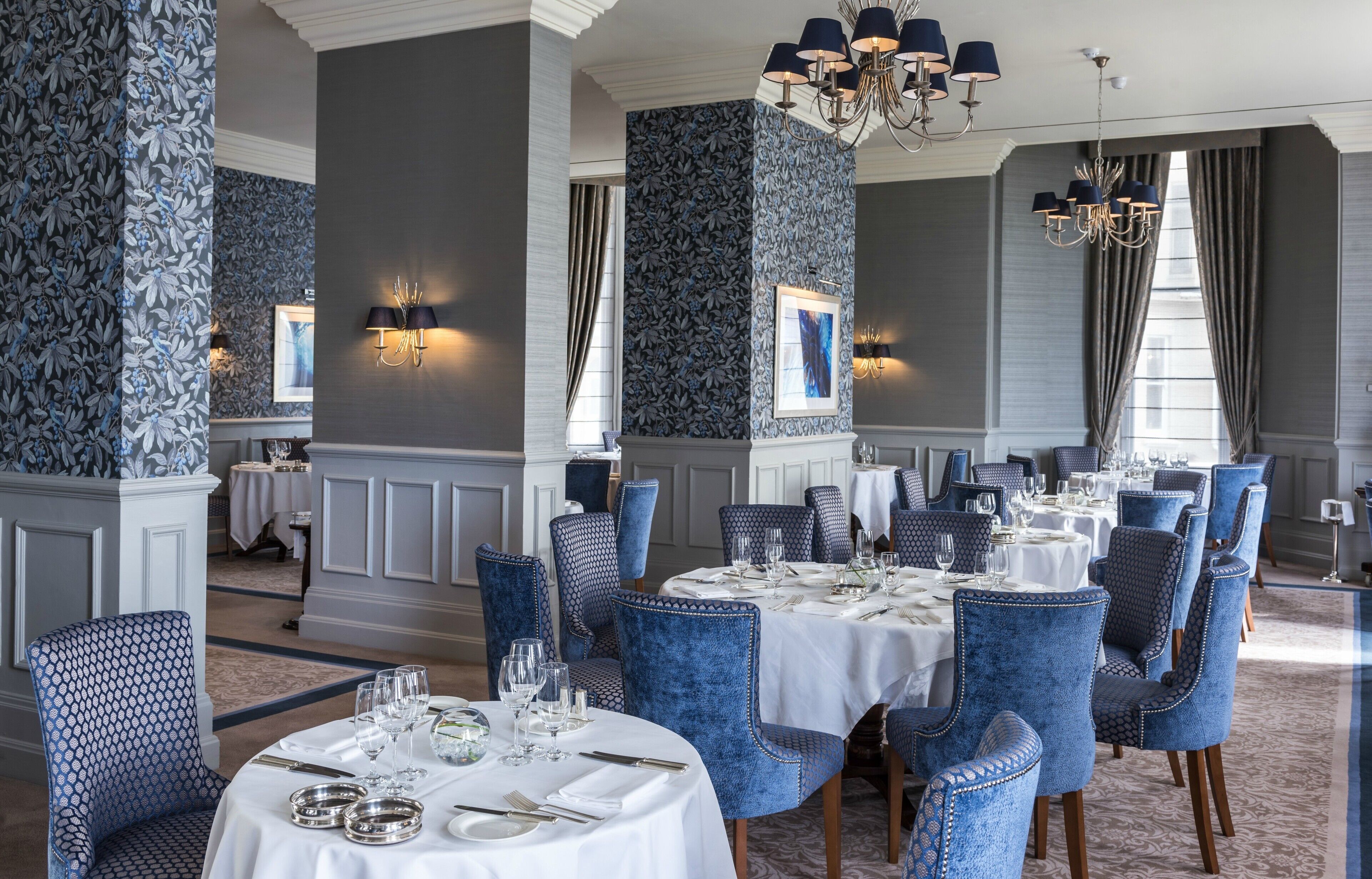A formal dining room with round tables, blue upholstered chairs, and elegant chandeliers.