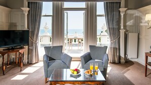 Une pièce dotée d’une grande fenêtre, de deux fauteuils rembourrés, d’une table basse avec des fruits et des boissons, et d’un téléviseur posé sur un meuble en bois.