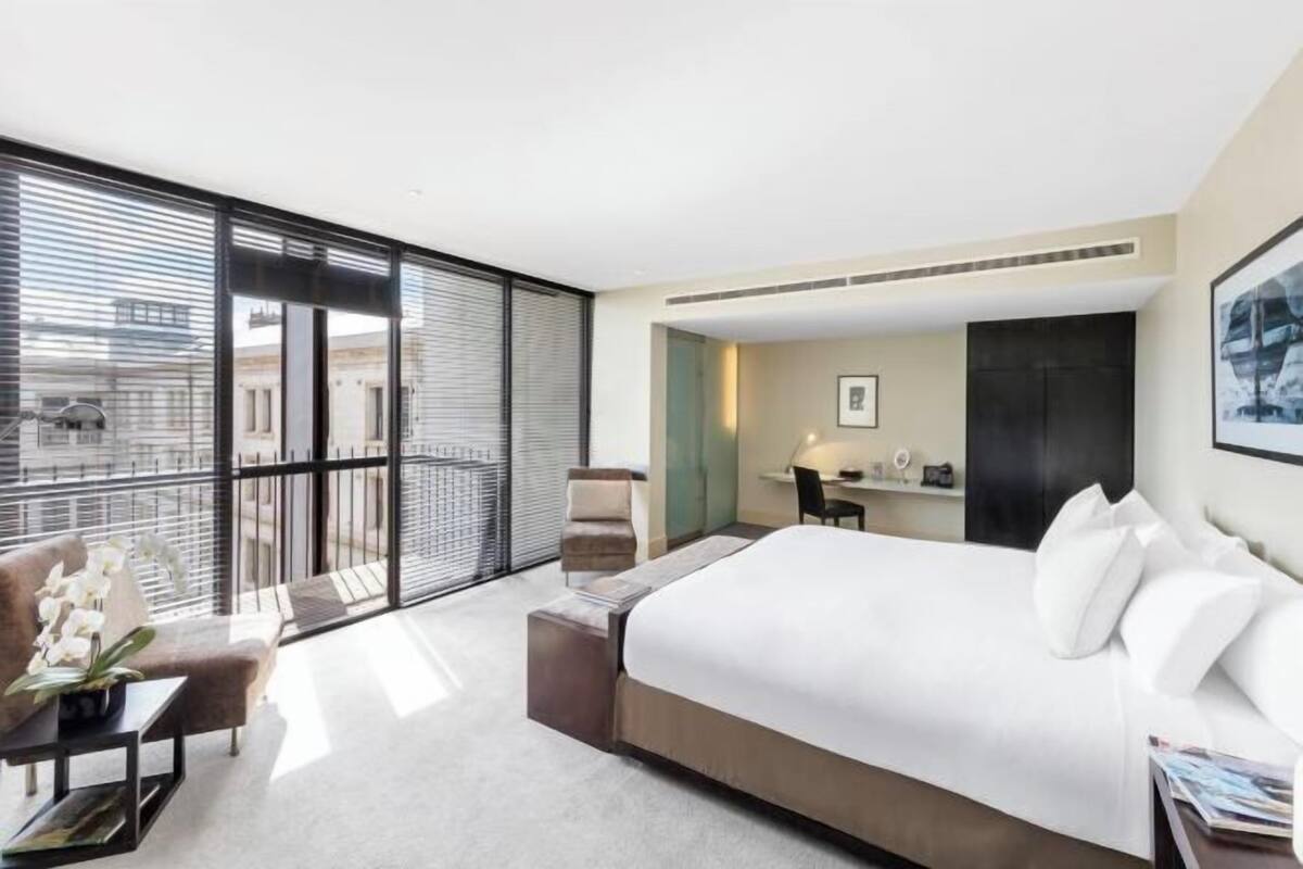Una habitación de hotel moderna con una cama grande, una zona de estar con una silla y una mesa, un escritorio con una silla, y vistas amplias por grandes ventanales.
