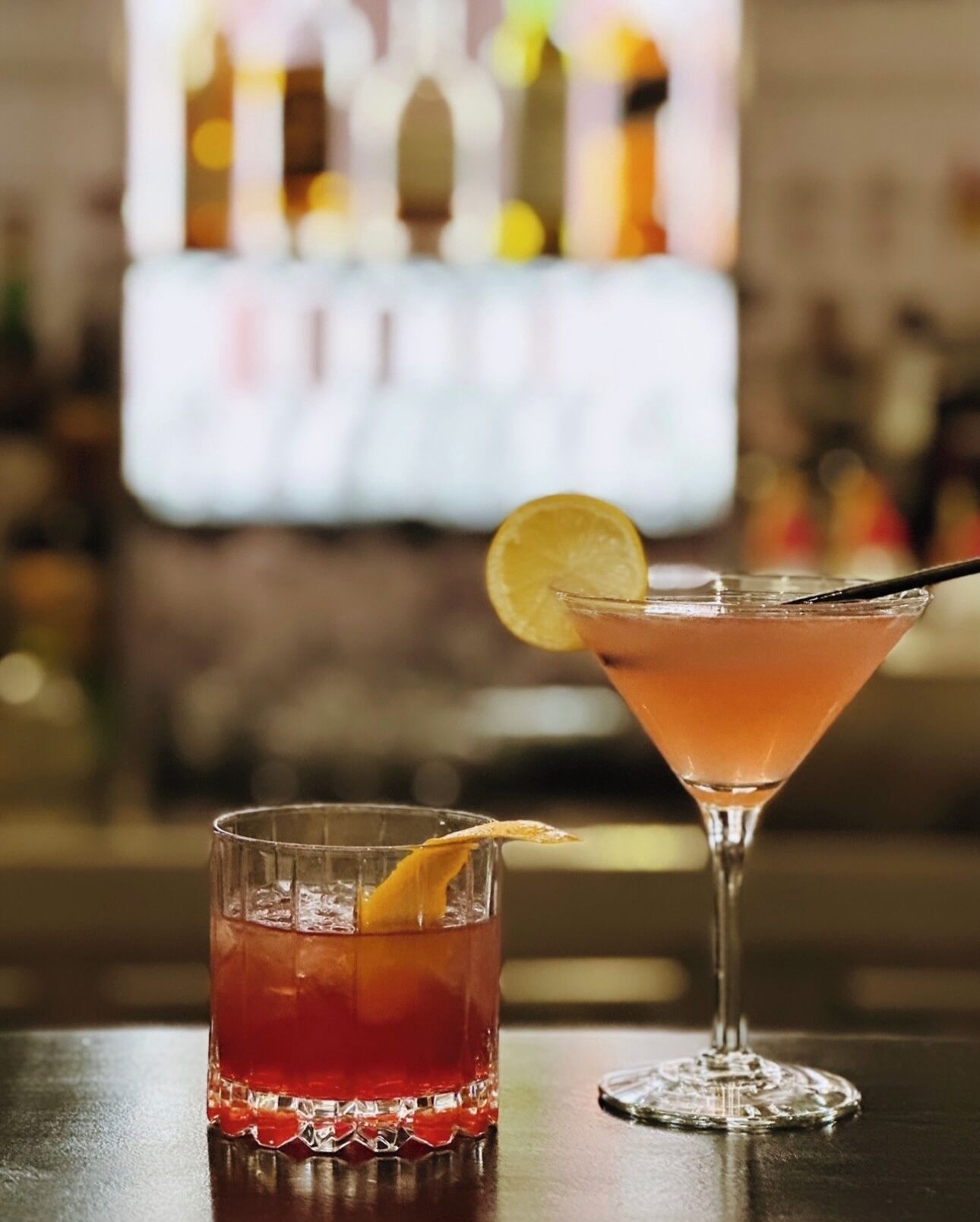 Zwei Cocktails auf einer Bartheke, einer im Kurzglas und einer im Martini-Glas, mit unscharfem Hintergrund, der eine Bar andeutet.