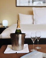 A hotel room with a bed, a nightstand with a lamp, a table with a champagne bottle and two glasses.