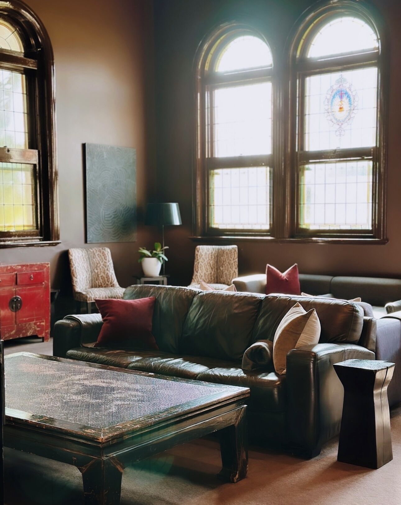 A cozy living room with a leather sofa, a coffee table, and a red dresser. There are two large windows with arched tops and a decorative stained glass window.