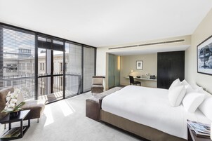 Quarto de hotel moderno com uma cama grande, área de estar com uma poltrona e uma mesinha, escrivaninha com cadeira e vista através de janelas do chão ao teto.