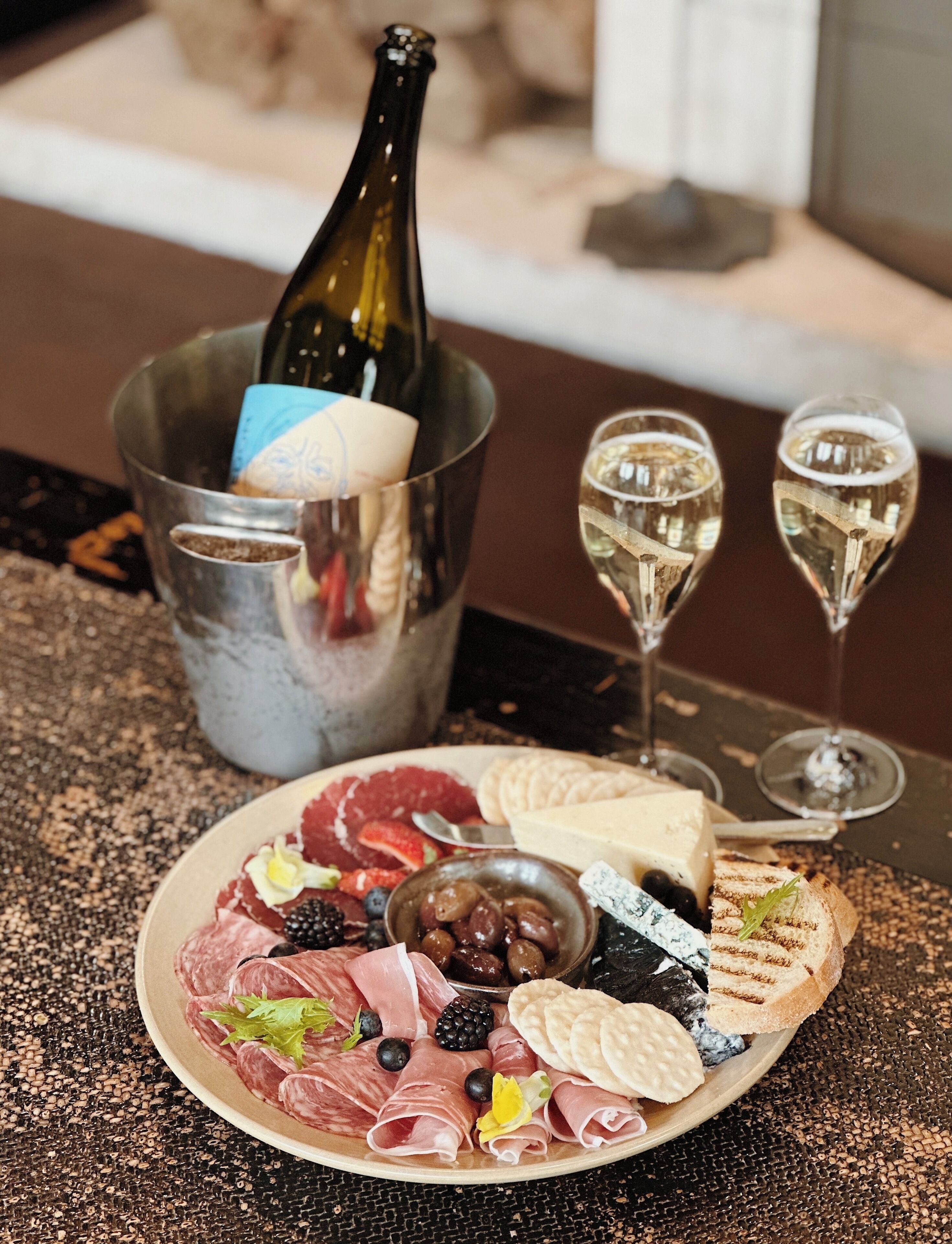 En anretningstallerken med forskellige slags pålæg, oste og frugter, samt to glas hvidvin og en flaske champagne i en metalspand.