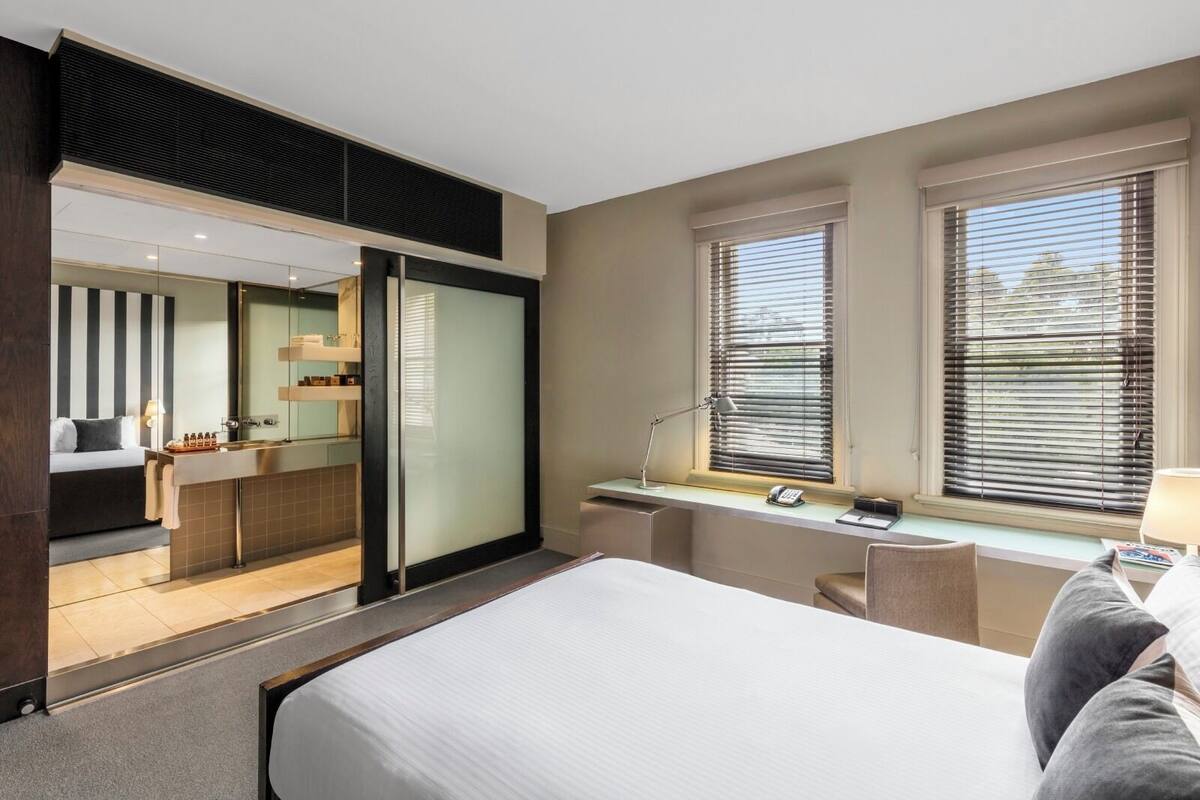 Habitación de hotel con una cama, un escritorio con lámpara, una silla y un ventanal con persianas.