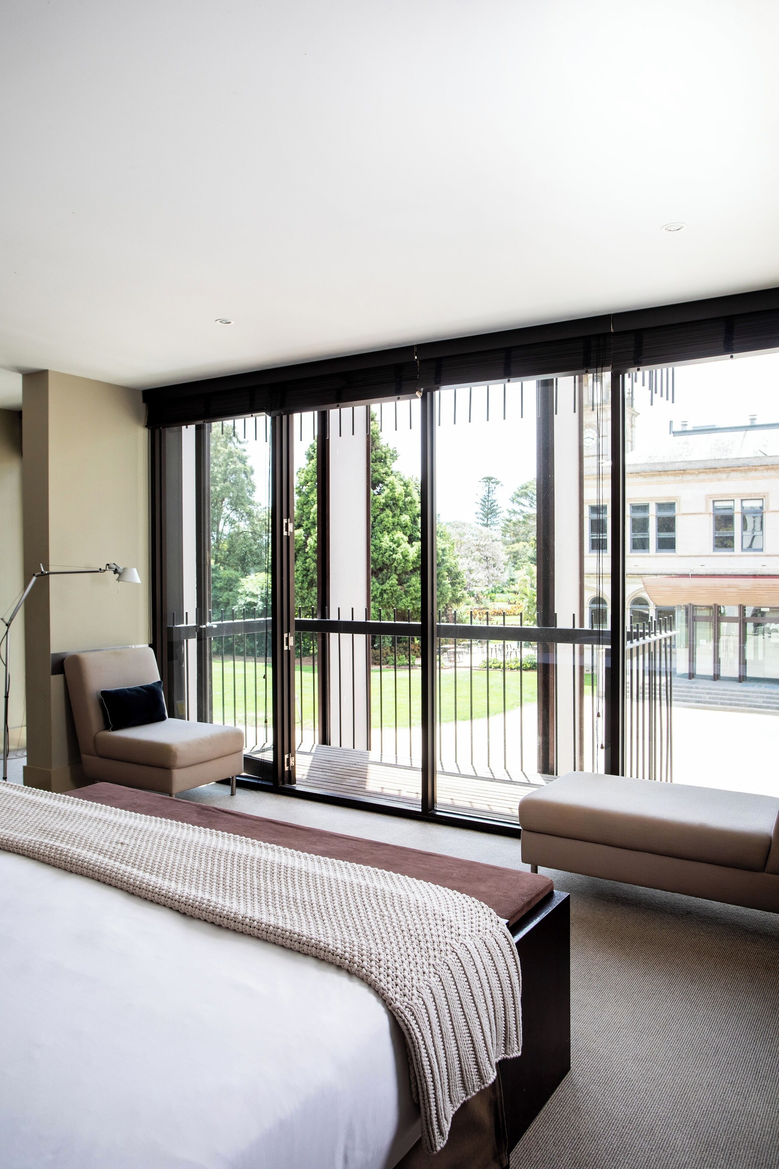 A modern hotel room with a large bed, a sofa, a chair, and a view of a courtyard through floor-to-ceiling windows.