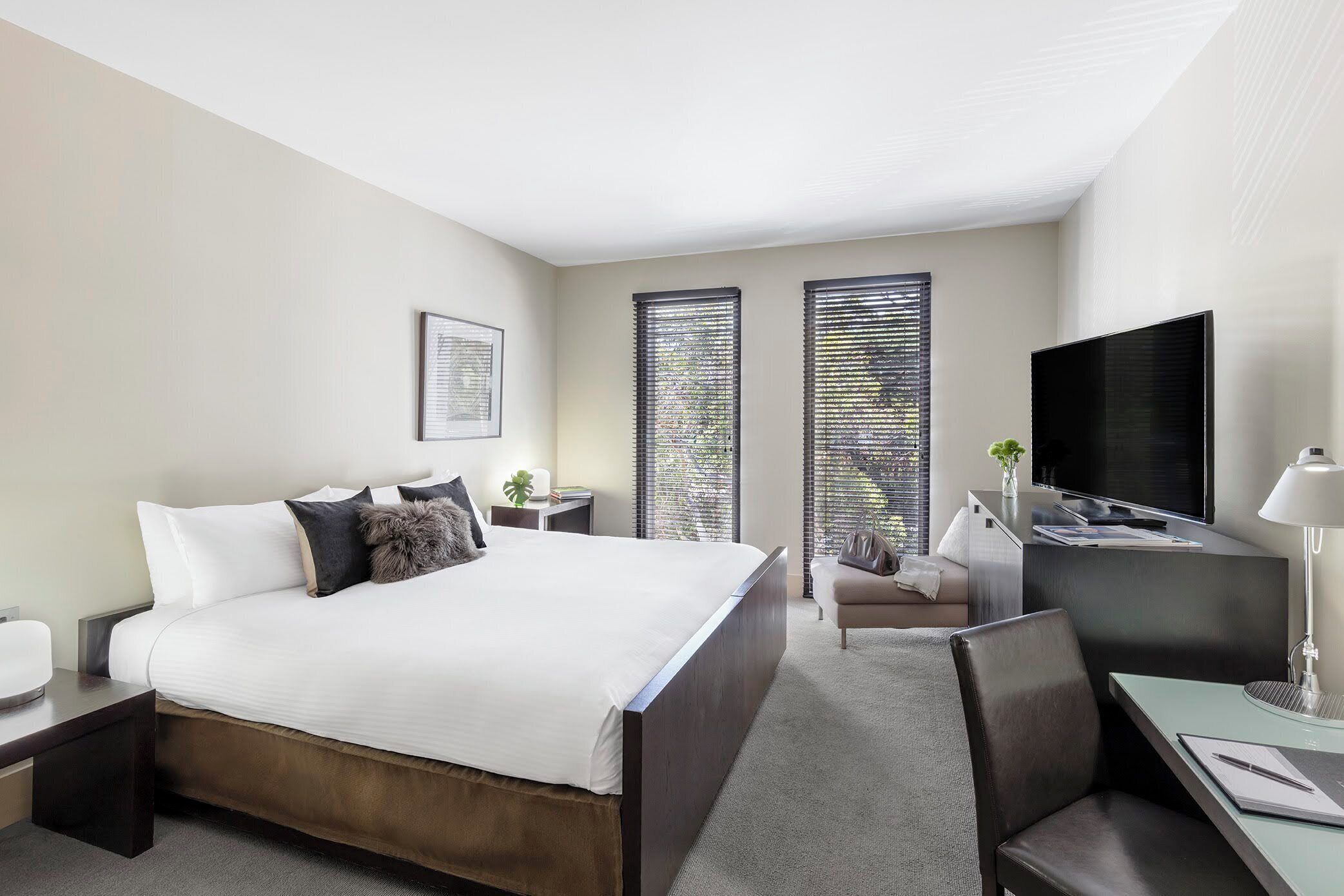 A modern hotel room with a large bed, a desk, a television, and two windows with blinds.