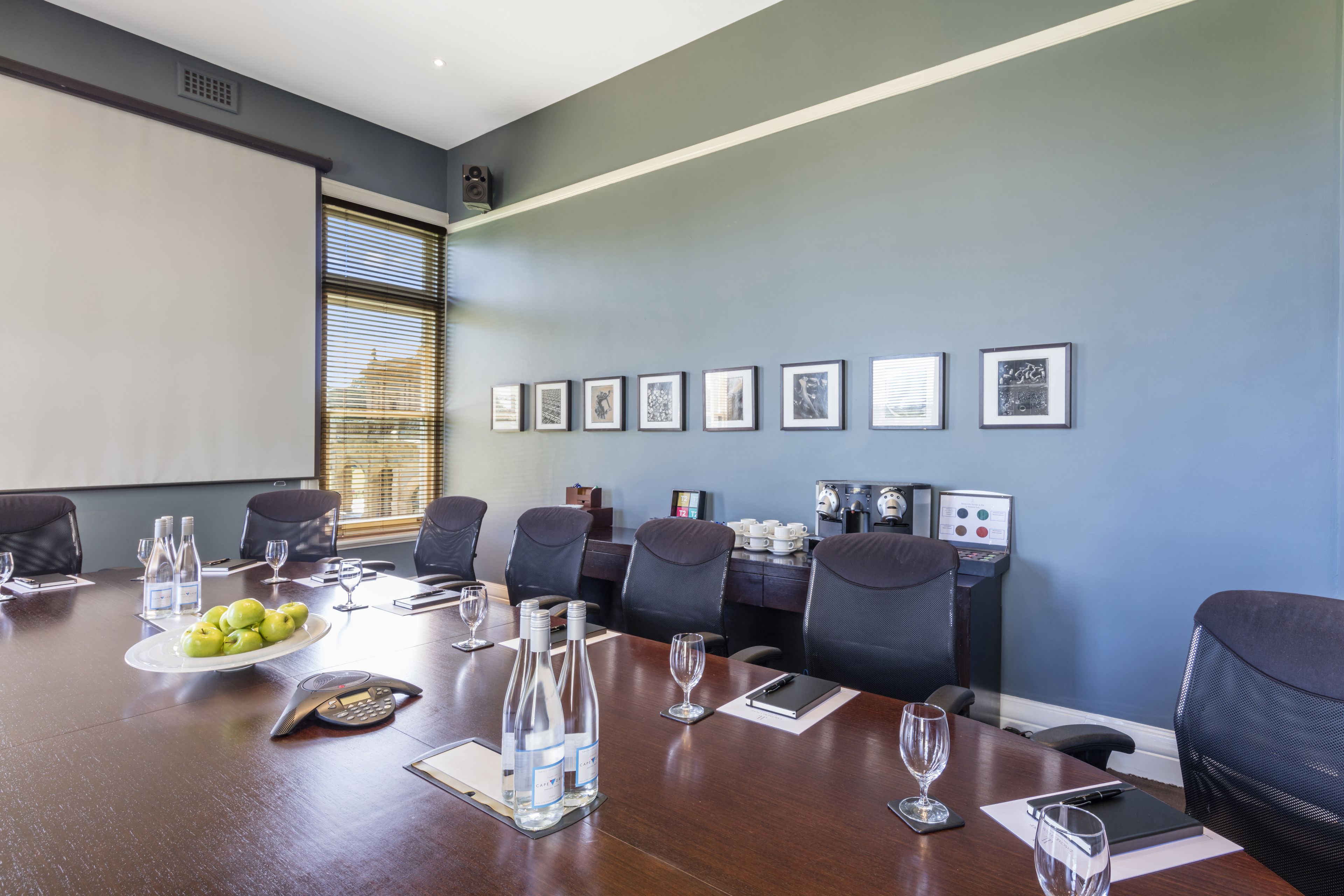 Ein Konferenzraum mit langem Tisch, Stühlen, einer Projektionsleinwand, gerahmten Bildern an der Wand und einer Kaffeemaschine.