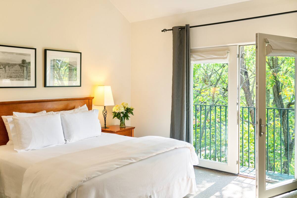 A hotel room with a large bed, a nightstand with a lamp and flowers, and a balcony with a view of trees.