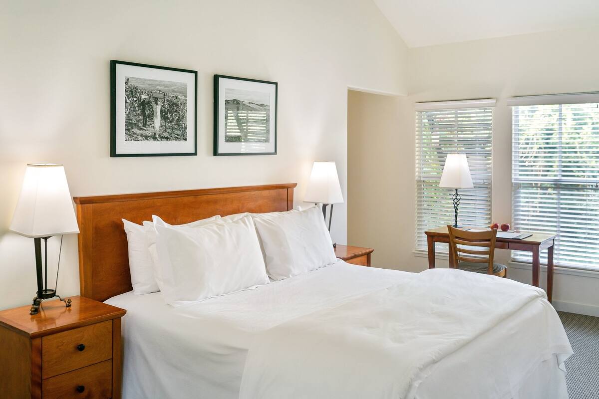A neatly made bed with white linens, two framed pictures on the wall, a wooden nightstand with a lamp, and a desk with a chair.